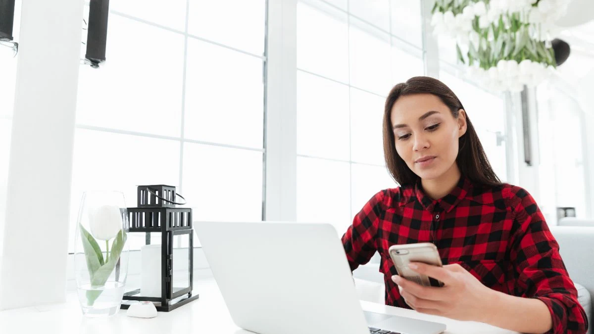 SDR in a red plaid shirt checking her phone at her laptop