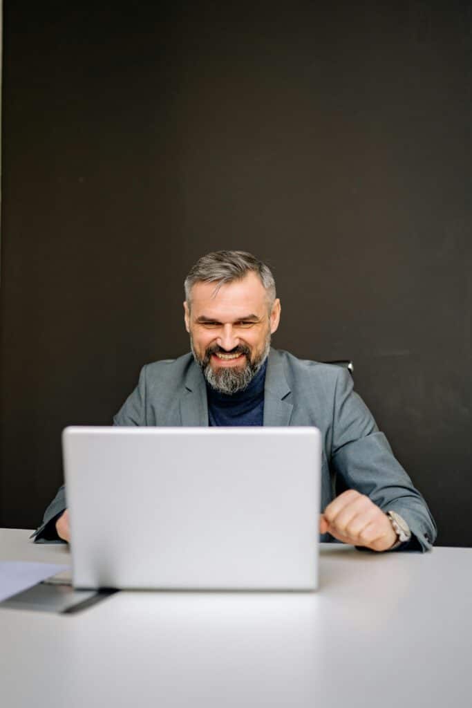 Smiling businessman in a gray suit using a laptop at a desk in an office setting.